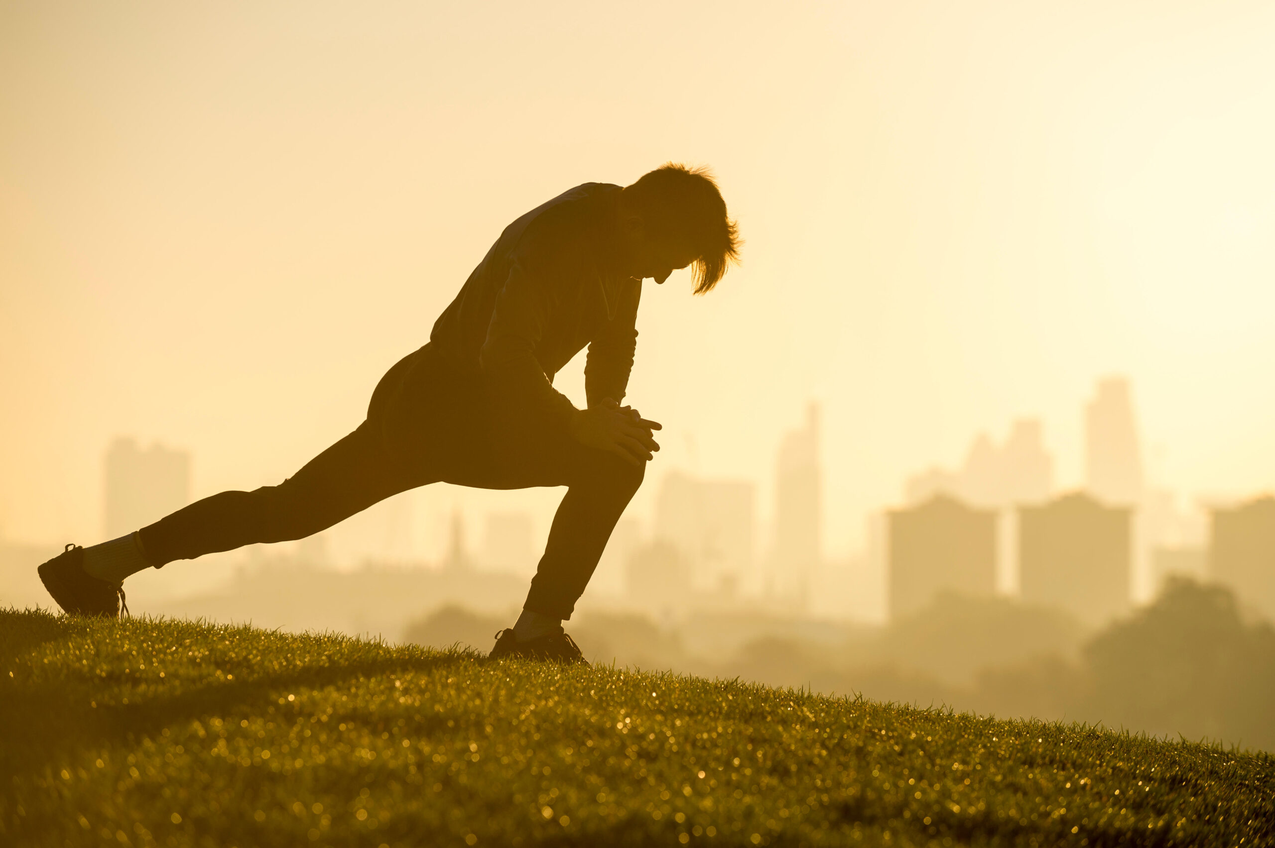 Male jogger stretching on the grassy top of Primrose Hill in front of a misty golden sunrise view of the London city skyline